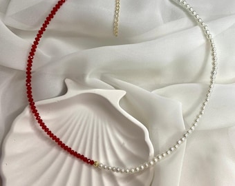 May include: A necklace with a half-and-half design. One side features small, red beads, while the other side has white pearls. The necklace has a gold-colored clasp and chain. The jewelry is displayed on a white seashell-shaped dish.