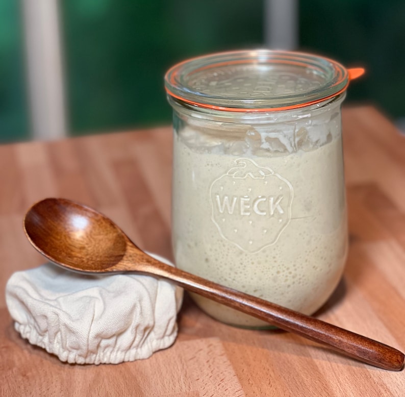 Pu&ograve; includere: Un barattolo di vetro con un coperchio bianco e una guarnizione di gomma. Il barattolo &egrave; pieno di un liquido chiaro e ha la scritta "WECK" stampata sul lato. Nell'immagine sono presenti anche un cucchiaio di legno e un panno bianco.