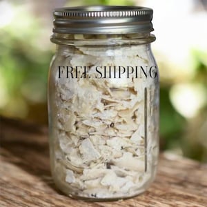 Puede incluir: Un tarro de cristal transparente con tapa plateada lleno de material claro y escamoso. El texto "FREE SHIPPING" está impreso en el tarro. El tarro está sobre una superficie de madera.