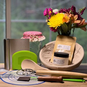 May include: A sourdough starter kit with a variety of tools and ingredients, including a wooden spoon, a dough scraper, a proofing basket, a jar of sourdough starter, a bag of sourdough starter, and a measuring tape. The kit also includes a bouquet of flowers in a vase.