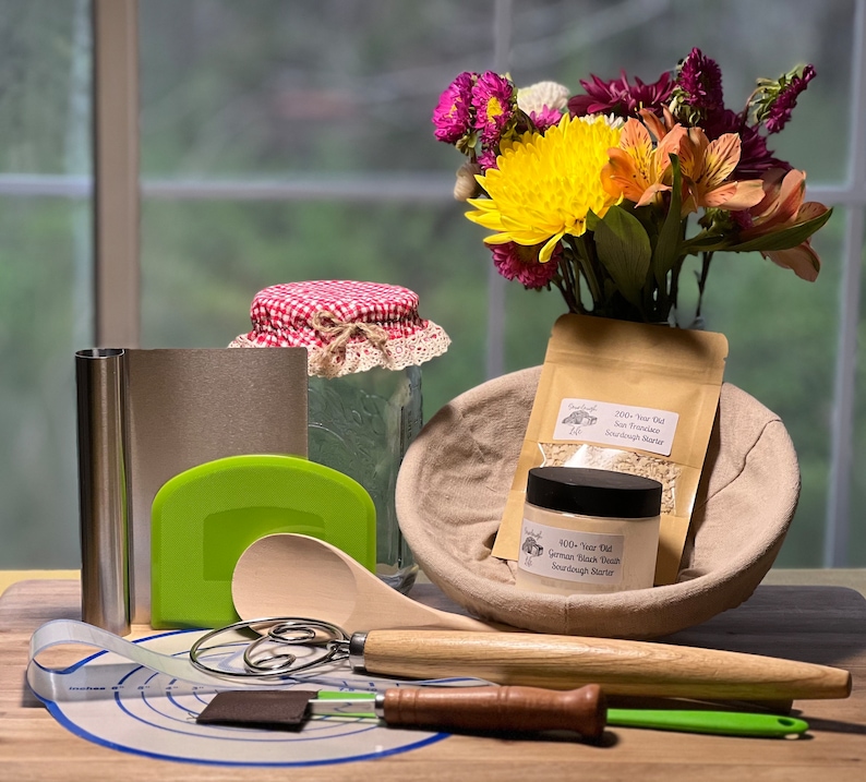 May include: A sourdough starter kit with a variety of tools and ingredients, including a wooden spoon, a dough scraper, a proofing basket, a jar of sourdough starter, a bag of sourdough starter, and a measuring tape. The kit also includes a bouquet of flowers in a vase.