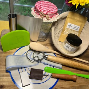 May include: A collection of sourdough starter baking supplies including a green dough scraper, a wooden spoon, a dough whisk, a green silicone spatula, a small wooden tool, a white ceramic rectangle, a brown leather rectangle, a blue and white silicone baking mat, a clear glass jar with a red and white checkered lid, a brown cloth bag with a white paper bag inside containing a white powder, and a small glass jar with a black lid containing a white powder.