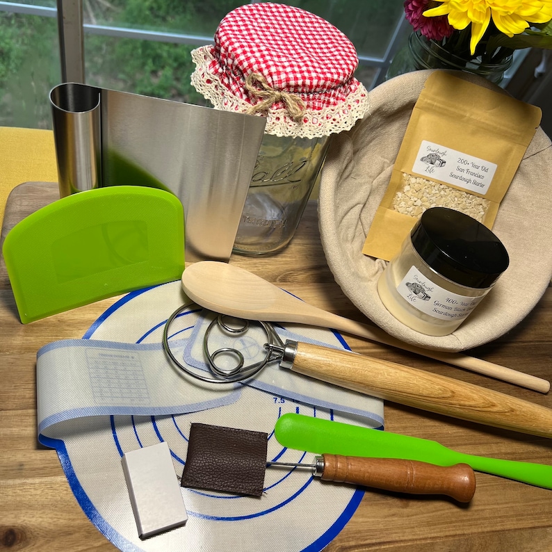 May include: A collection of sourdough starter baking supplies including a green dough scraper, a wooden spoon, a dough whisk, a green silicone spatula, a small wooden tool, a white ceramic rectangle, a brown leather rectangle, a blue and white silicone baking mat, a clear glass jar with a red and white checkered lid, a brown cloth bag with a white paper bag inside containing a white powder, and a small glass jar with a black lid containing a white powder.