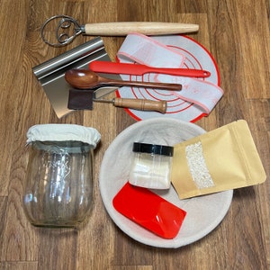 May include: A set of baking tools and supplies including a wooden dough whisk, a metal scraper, a glass jar, a red silicone spatula, a wooden spoon, a white bowl, a bag of flour, and a small container of yeast.