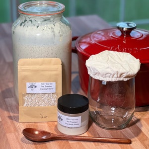 Pu&ograve; includere: Un kit di lievito madre. Il kit include un barattolo di vetro con un coperchio di stoffa bianca, un barattolo di vetro pi&ugrave; piccolo con un coperchio nero, una busta di carta marrone con un'etichetta che recita "200+ Year Old San Francisco Sourdough Starter" e un cucchiaio di legno. Il kit &egrave; su una superficie di legno.