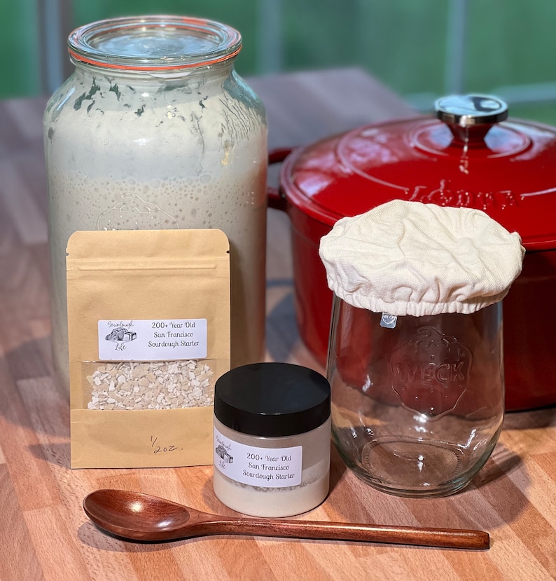 Pu&ograve; includere: Un kit di lievito madre. Il kit include un barattolo di vetro con un coperchio di stoffa bianca, un barattolo di vetro pi&ugrave; piccolo con un coperchio nero, una busta di carta marrone con un'etichetta che recita "200+ Year Old San Francisco Sourdough Starter" e un cucchiaio di legno. Il kit &egrave; su una superficie di legno.