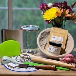 May include: A collection of sourdough starter supplies, including a clear glass jar with a lid, a white bowl with a cloth liner, a wooden rolling pin, a dough scraper, a metal whisk, a green silicone spatula, a small white box, and two jars of sourdough starter. The jars are labeled "2000+ Year Old San Francisco Sourdough Starter" and "400+ Year Old German Black Rye Sourdough Starter".