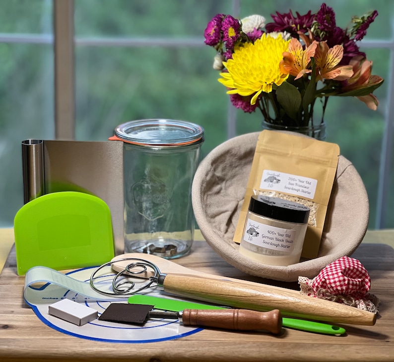 May include: A collection of sourdough starter supplies, including a clear glass jar with a lid, a white bowl with a cloth liner, a wooden rolling pin, a dough scraper, a metal whisk, a green silicone spatula, a small white box, and two jars of sourdough starter. The jars are labeled "2000+ Year Old San Francisco Sourdough Starter" and "400+ Year Old German Black Rye Sourdough Starter".