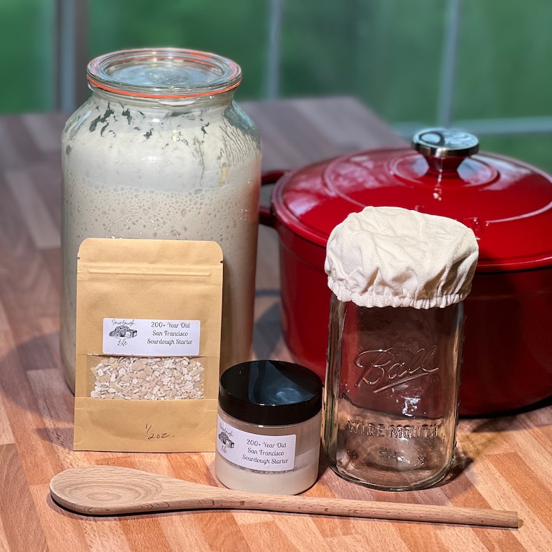 Pu&ograve; includere: Un primo piano di un kit di lievito madre. Il kit include un barattolo di vetro con un coperchio in tessuto bianco, un barattolo di vetro pi&ugrave; piccolo con un coperchio nero, una busta di carta marrone con un'etichetta che recita "200+ Year Old San Francisco Sourdough Starter 1/2 oz", e un cucchiaio di legno.