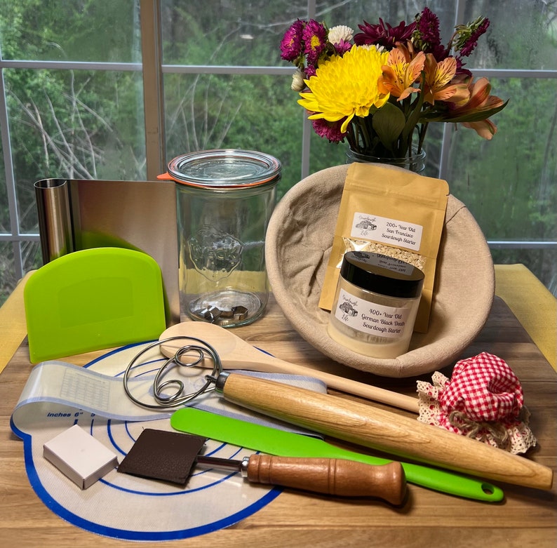 May include: A wooden cutting board with a variety of baking tools, including a dough whisk, a green spatula, a brown leather scraper, a clear glass jar, a white bowl, and two bags of sourdough starter. The bags are labeled "250+ Year Old San Francisco Sourdough Starter" and "400+ Year Old German Black Forest Sourdough Starter".