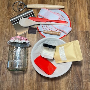 May include: A baking starter kit with various red and wooden tools, including a dough scraper, whisk, and silicone spatula.  A beige bread proofing basket, a mason jar, and small container of starter are also included.  The kit is arranged on a wood surface.