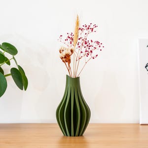 Peut inclure: Un vase en céramique vert avec un design unique et côtelé. Le vase est rempli de fleurs séchées, y compris de petites fleurs blanches et de longues tiges fines avec des baies rouges.