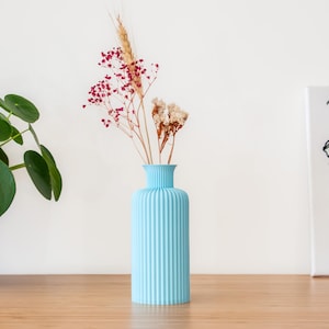 Peut inclure: Un vase en céramique bleu clair avec des nervures verticales, contenant des fleurs séchées. Le vase est posé sur une surface en bois.