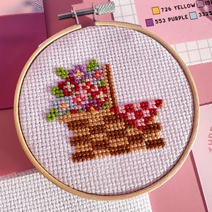 May include: A cross-stitch embroidery project in a wooden hoop. The design shows a pixelated basket of flowers in red, purple, and green. The background includes pink paper, embroidery floss, and a needle card.