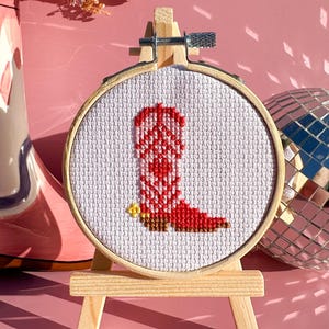 May include: A cross-stitch embroidery of a red and white cowboy boot in a wooden hoop, displayed on a small easel. The background is a solid pink, with a disco ball and a vase of dried flowers. The boot has a yellow spur.