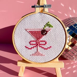 May include: A cross-stitch embroidery of a pink cocktail in a martini glass, with a bow, olive, and mint leaf, set in a wooden hoop. The embroidery is displayed on a small wooden easel, with a disco ball in the background.