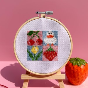 Puede incluir: Un pequeño bastidor de bordado de punto de cruz redondo con un diseño colorido de cerezas, un pato, una flor y una fresa. El bordado se exhibe en un pequeño caballete de madera, con una fresa de cerámica cerca.