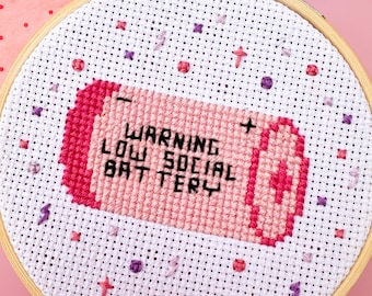 May include: A pink and white cross-stitch embroidery hoop with the text "WARNING LOW SOCIAL BATTERY" in black lettering. The embroidery is of a pink battery with a plus sign on the top.