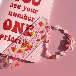 Op de afbeelding: Een roze en witte kralen armband met een roze strik bedel en een transparante bladwijzer met een aardbei en roze strik patroon. De bladwijzer heeft de tekst "You are your number one priority" op een roze achtergrond.