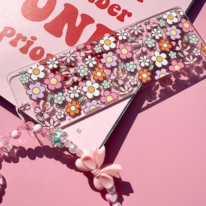 May include: A clear ruler decorated with colorful flowers and bows. The ruler features pink, orange, yellow, and white flowers. A beaded charm with a pink bow and heart-shaped beads is attached. The background is pink, with text that reads "ONE".