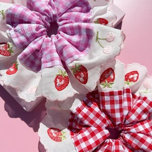 Könnte beinhalten: Zwei Stoffhaargummis mit Karomuster und gestickten Erdbeeren. Ein Haargummi ist in rosa und weißem Karomuster, das andere in rot und weißem Karomuster.