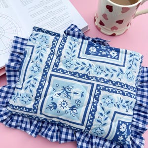 May include: A blue and white floral patterned quilted pillow with a ruffled gingham border. The pillow is next to an open book titled "Navigating a birth chart" and a mug with heart designs.