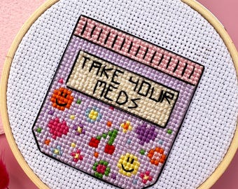 May include: A purple and pink cross-stitch embroidery hoop with the text "Take Your Meds" on a pill bottle design. The bottle is filled with colorful hearts, flowers, and smiley faces.