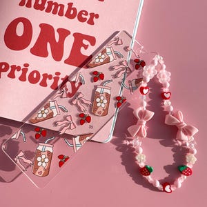 Op de afbeelding: Een roze en witte kralenketting met roze strikken, aardbeienbedels en witte bloemenbedels. De ketting ligt op een roze achtergrond met een transparante acryl boekenlegger met een aardbeienmelkpatroon.