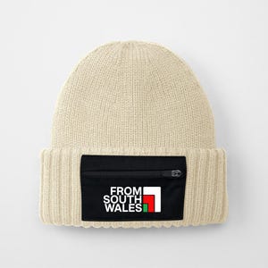 May include: A beige knitted beanie with a black zippered pocket on the front. The pocket has a white logo that says "FROM SOUTH WALES" with a red and green flag design.