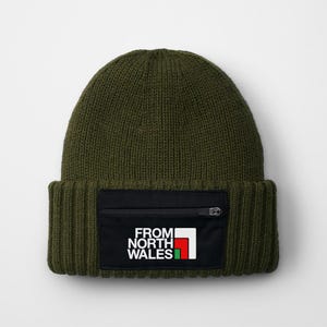 May include: A green knitted beanie with a black zipped pocket on the front. The pocket has a white logo that says "From North Wales" with a red and green flag design.