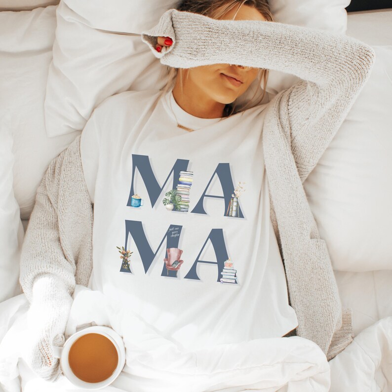 May include: A white t-shirt with the word 'MAMA' printed in blue letters. Each letter is decorated with small illustrations of books, plants, and other home decor items. The t-shirt is worn by a person lying in bed.