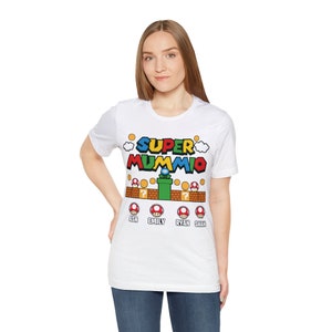 May include: White t-shirt featuring a colourful graphic of a video game character and the text "SUPER MUMMIO". The graphic includes a green pipe, red mushrooms, and gold coins. The text "ASH", "EMILY", "RYAN", and "SHAN" is printed below the graphic.