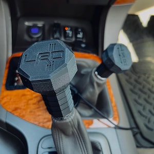 May include: Close-up of a black gear shift knob with a raised "LRD" logo. The knob is attached to a leather-wrapped gear shift lever in a car.