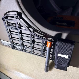 Puede incluir: Panel Molle de metal negro con el texto "170 M" y "170 Molle" instalado en un vehículo. Una linterna negra con la parte superior naranja y una bolsa negra con un parche blanco están adjuntas.