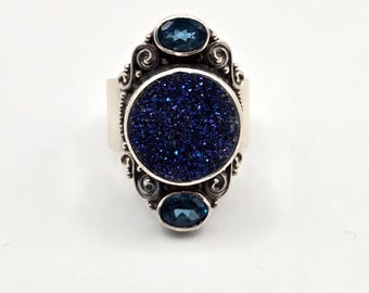 May include: A silver ring with a blue sparkly stone and two smaller blue stones. The ring has a detailed design.