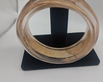 Könnte beinhalten: Ein transparentes, durchsichtiges Armreif-Armband mit einem braunen Marmormuster. Der Armreif wird auf einem schwarzen Samtständer präsentiert.