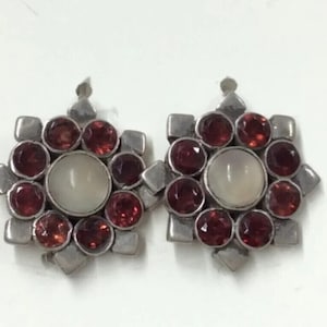 May include: A pair of silver earrings with a starburst design. Each earring features a central white stone surrounded by eight red gemstones.