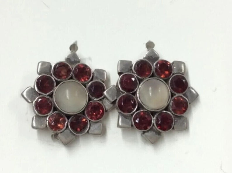 May include: A pair of silver earrings with a starburst design. Each earring features a central white stone surrounded by eight red gemstones.