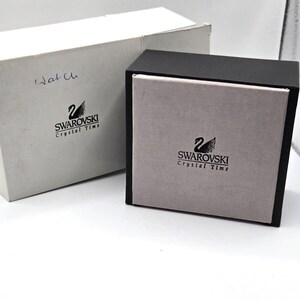 May include: Two boxes for a Swarovski Crystal Time watch. The smaller box is black with a gray interior and the larger box is white with the Swarovski logo and the words "Crystal Time".