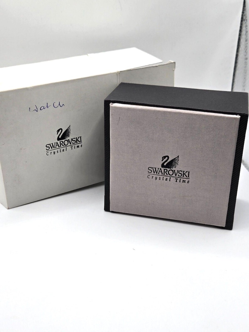 May include: Two boxes for a Swarovski Crystal Time watch. The smaller box is black with a gray interior and the larger box is white with the Swarovski logo and the words "Crystal Time".
