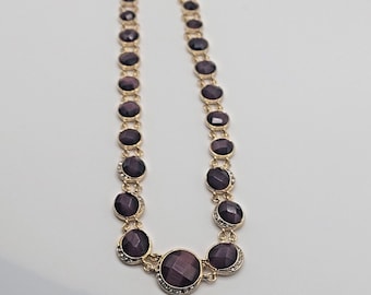 May include: A gold necklace with a chain of round, faceted, dark purple stones. The stones are set in gold and have a small row of clear rhinestones around the edge.