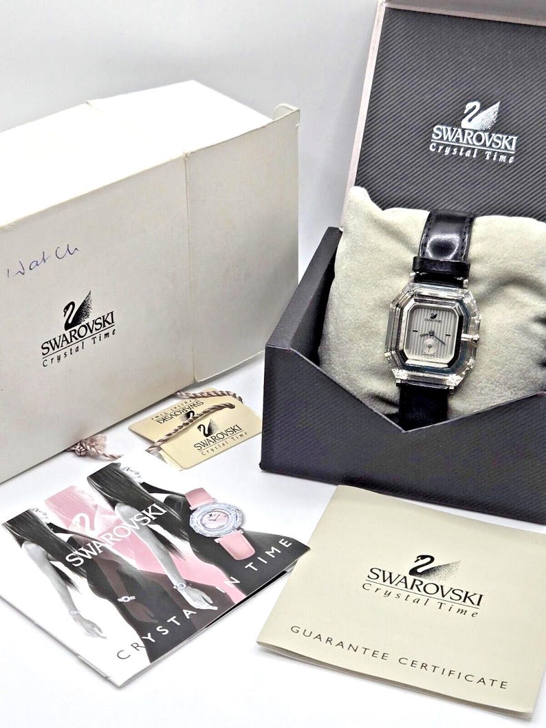 May include: A white box with the text "Swarovski Crystal Time" and a swan logo. A black box with the text "Swarovski Crystal Time" and a swan logo. A white and pink brochure with the text "Swarovski Crystal Time". A black and white guarantee certificate with the text "Swarovski Crystal Time" and "Guarantee Certificate". A silver watch with a black leather band and a crystal face. The watch is in a black box with a white pillow.