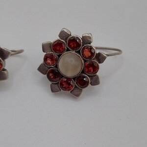 May include: A pair of silver earrings with a flower design. The earrings have a white center stone surrounded by red stones.