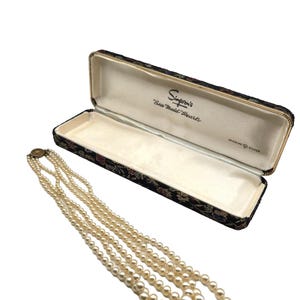 May include: A triple-strand pearl necklace with a gold clasp, next to an open black jewellery box with floral detailing. The box is labelled "Simpson's Sea Maid Pearls."