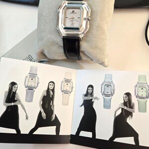 May include: A white watch with a crystal face and black leather band. The watch is displayed on a white cushion. The image also shows four women wearing black dresses and different colored watches.