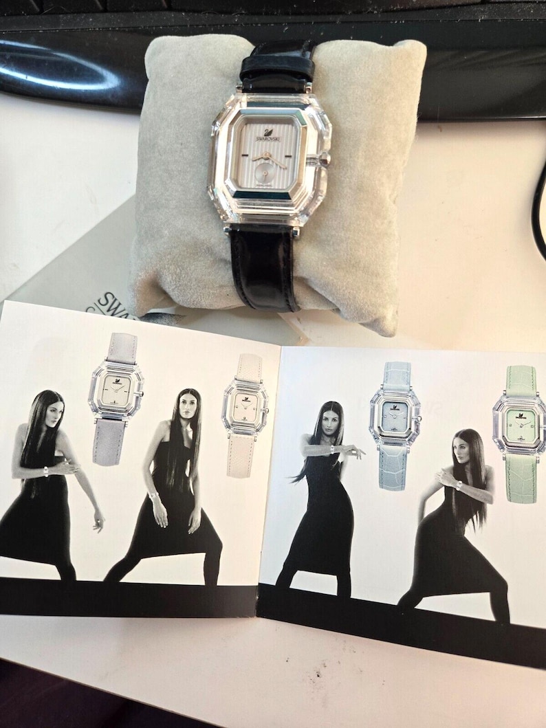 May include: A white watch with a crystal face and black leather band. The watch is displayed on a white cushion. The image also shows four women wearing black dresses and different colored watches.