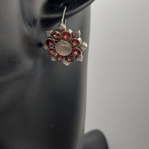 May include: Silver earrings with a circular design featuring a white center stone surrounded by eight red gemstones.