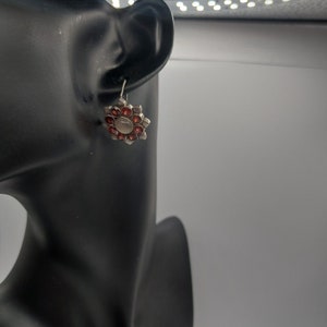 May include: Silver dangle earrings with a red flower design. The flower has a central white stone surrounded by smaller red stones.