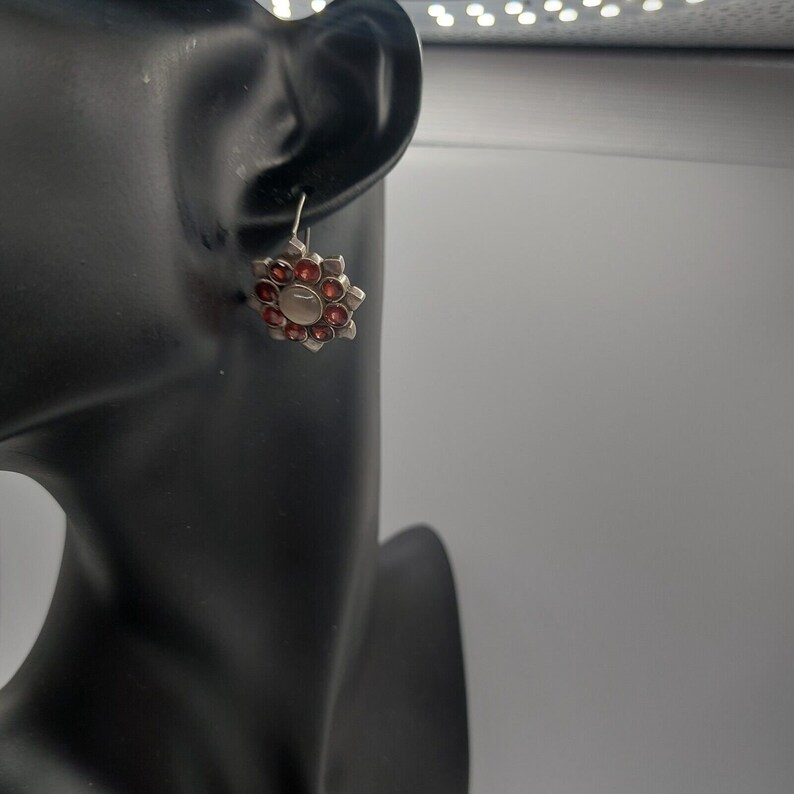 May include: Silver dangle earrings with a red flower design. The flower has a central white stone surrounded by smaller red stones.