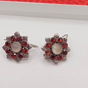 May include: A pair of silver earrings with a starburst design. Each earring features a central white pearl surrounded by 12 red gemstones.